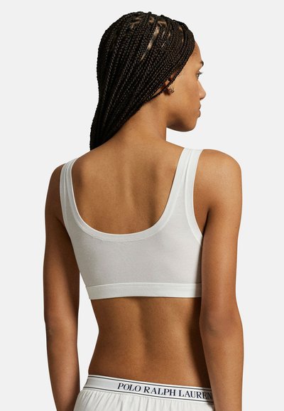 Γυναίκα με πλεξούδες, φοράει λευκό αθλητικό σουτιέν και λευκά σορτς Polo Ralph Lauren, απεικονίζεται από πίσω σε απλό φόντο.