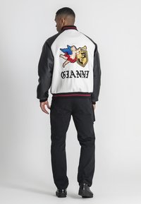 Mand iført sort og hvid varsity-jakke med farverig engel og "GIANNI" tekst på ryggen, sorte bukser og sorte sko, vendt væk.