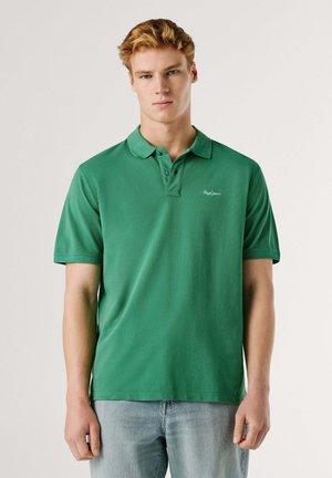 Joven de piel clara y cabello rubio, vistiendo una camiseta polo verde de manga corta y jeans azul claro, de pie frente a un fondo liso.