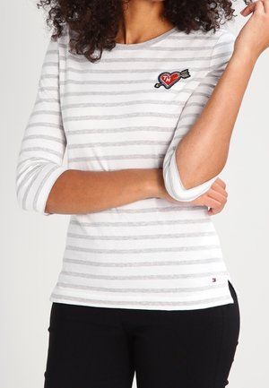 Žena v dlouhém rukávu bílého a světle šedého pruhovaného trička s nášivkou srdce a šípu na hrudi, doplněného černými kalhotami.