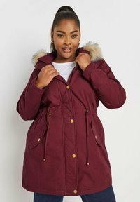 Burgundy parka takki, jossa on tekoturkishuppu, kiristettävä vyötärö ja kultaiset metallinapit. Sisältää kaksi etutaskua ja tikatun suunnittelun.