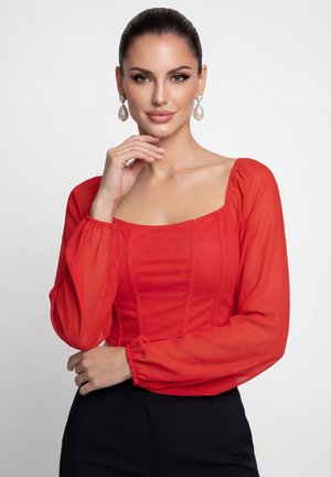 Žena s tmavými vlasy uhlazenými dozadu, která má na sobě červený top s dlouhými rukávy a perlové visací náušnice, pózuje s jednou rukou blízko brady.