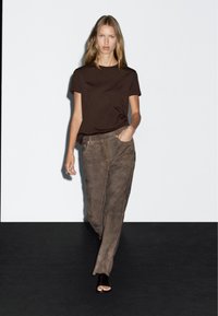 Brun kortærmet skjorte parret med lysebrune ruskindsbukser. Outfittet fremhæver bløde teksturer og minimalistisk design. Sort gulv baggrund.