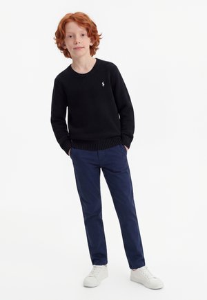 Garçon aux cheveux roux portant un pull noir, un pantalon bleu marine et des baskets blanches, debout les mains dans les poches sur fond blanc.