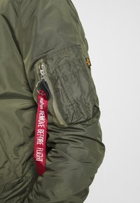 Giacca bomber verde oliva con tasca con zip sul braccio, caratterizzata da un'etichetta rossa "RIMUOVERE PRIMA DEL VOLO" e un accento di logo giallo. Tessuto dalla superficie liscia.