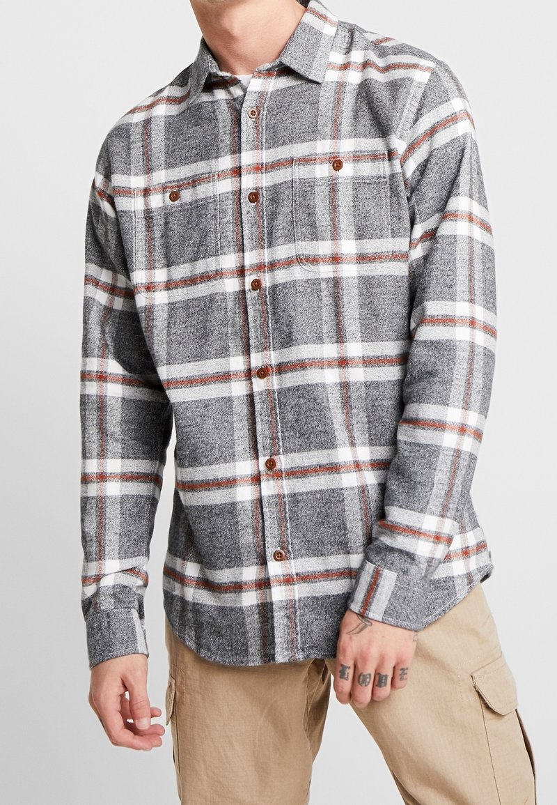 Camisa de franela a cuadros en gris y blanco con acentos en naranja, cierre de botones, dos bolsillos en el pecho y mangas largas. Textura suave.