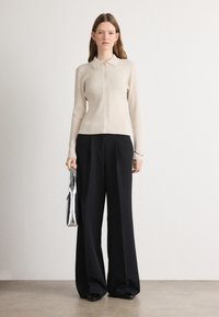 Beige ribbet cardigan med spiss krage og knappelukking, kombinert med vide svarte bukser og en sølvveske. Glatte teksturer.