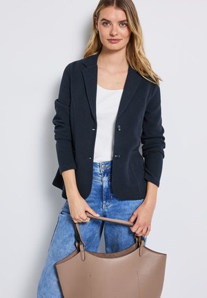 Woman wearing a dark blazer, white top, and blue jeans holding a large taupe leather handbag with both hands.