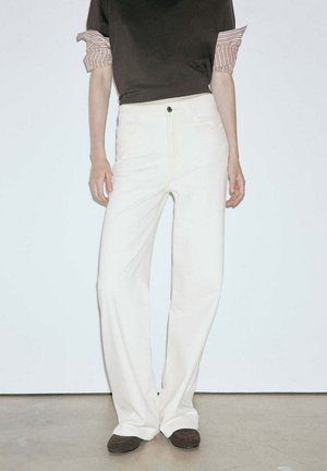 Personne portant un pantalon blanc taille haute à jambes larges, un haut sombre avec des manches rayées retroussées et des chaussures foncées, debout devant un fond clair uni.