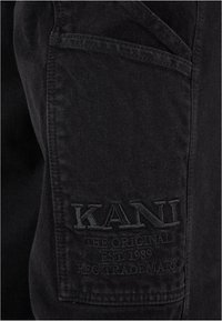 Pantaloni cargo neri in cotone con un tessuto testurizzato, una tasca laterale con il logo "KANI" in rilievo e dettagli di cucitura.