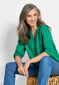 Blusa verde con bottoni davanti e maniche arrotolate, abbinata a jeans blu. Il tessuto appare liscio e leggero. Presente uno sgabello in rattan.