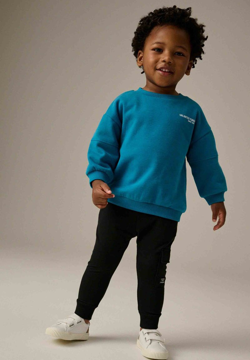 Bambino piccolo con felpa blu, pantaloni neri e scarpe da ginnastica bianche in piedi che sorride contro uno sfondo uniforme.