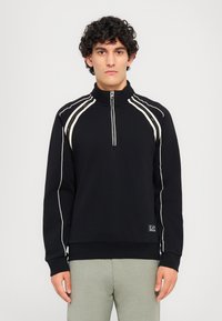 Svart høyhalset sweatshirt med hvite detaljer langs skuldrene og glidelås foran. Har ribbestrikkede mansjetter og et logo-patch på kanten.