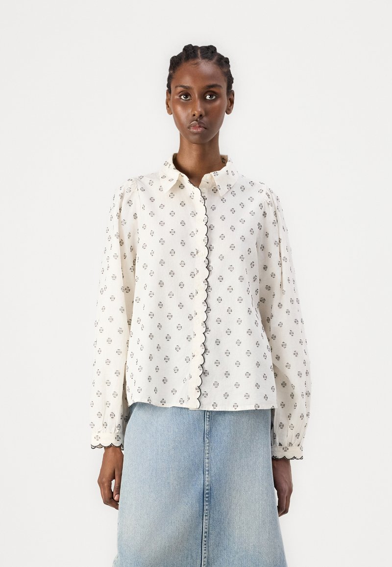 Vit blus med små svarta blommönster, scallopade kanter och knappstängning framtill. Långa puffärmar, bärs över en jeanskjol.