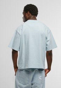 Tricou oversized, de culoare albastru deschis, cu mânecă scurtă, realizat dintr-un material moale. Are un tiv drept și un detaliu cu logo pe spate, combinat cu șorturi din denim deschis.