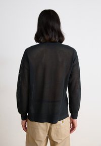 Black, knitted sweater with a textured open weave, ribbed cuffs, and hem. Viewed from the back, paired with beige pants featuring a logo.