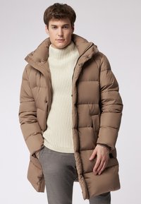 Cappotto puffer marrone con cappuccio, caratterizzato da un design trapuntato e bottoni a scatto. Indossato sopra un maglione a collo alto crema e pantaloni grigi.