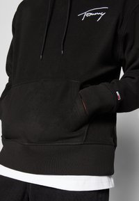 Svart fleece hoodie med en framficka. Har en vit broderad logotyp, dragsko och ribbade ärmslut och nederkant. Lags över en vit skjorta.
