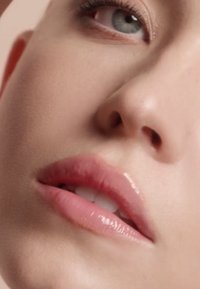 Primer plano de un rostro que muestra piel suave, labios rosados y voluminosos con acabado brillante, ojos de color claro y un maquillaje sutil con tonos naturales.