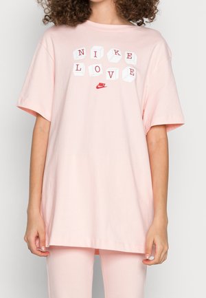 Personne portant un t-shirt rose clair avec des lettres en blocs 3D formant "NIKE LOVE" et un petit logo rouge Nike sous le texte.