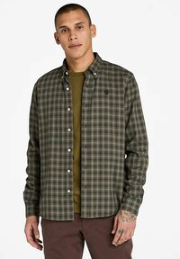 Uomo che indossa una camicia a quadri verde e marrone con bottoni sopra una maglietta verde oliva, abbinata a pantaloni marroni, con la mano sinistra tatuata visibile.