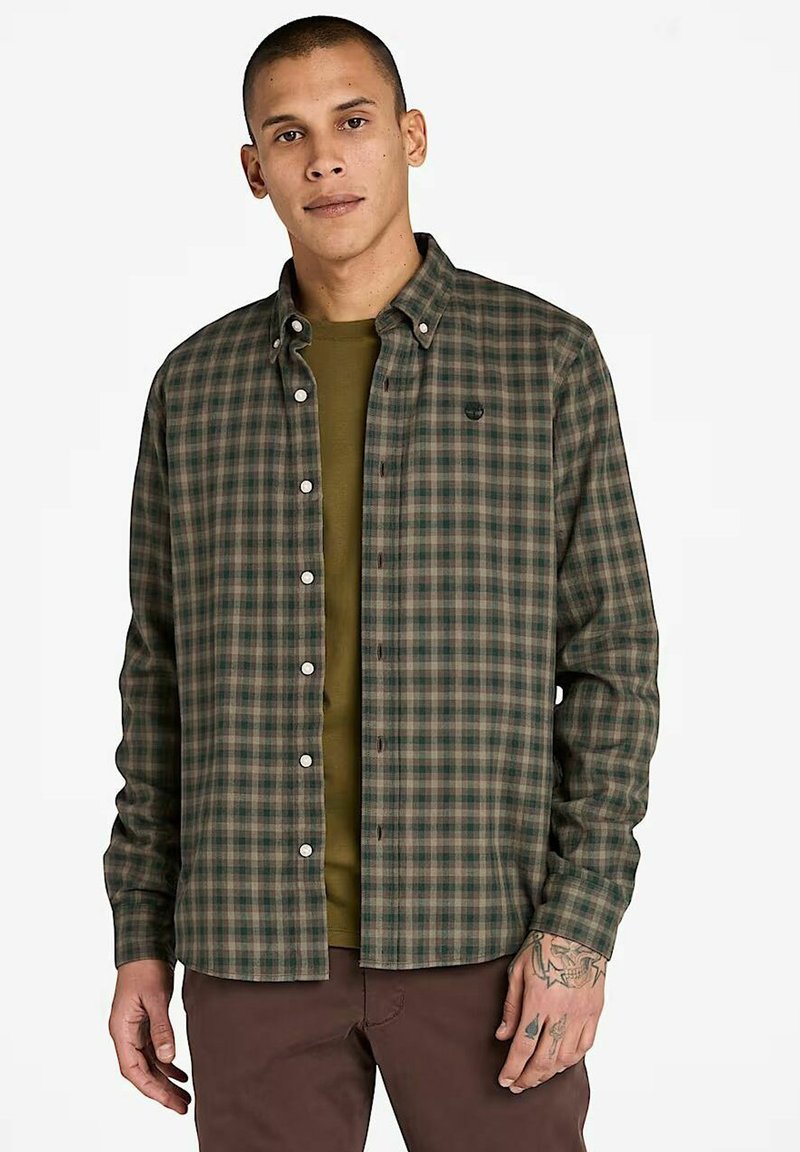 Uomo che indossa una camicia a quadri verde e marrone con bottoni sopra una maglietta verde oliva, abbinata a pantaloni marroni, con la mano sinistra tatuata visibile.