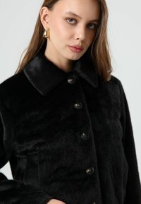 Veste en fausse fourrure noire avec col, dotée de grands boutons et d'une texture douce. Coupe cintrée avec une finition lisse et élégante.