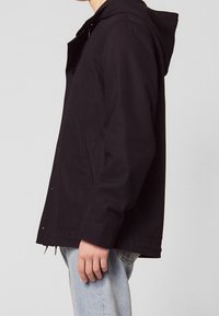 Czarna kurtka z kapturem wykonana z gładkiego materiału. Posiada zapięcie na guziki, boczne kieszenie oraz luźny krój.