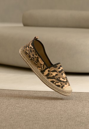 Espadrilio batai su leopardo rašto audiniu, juodais apvadais ir austu jutos padais, pakibę virš smėlio spalvos kilimo.