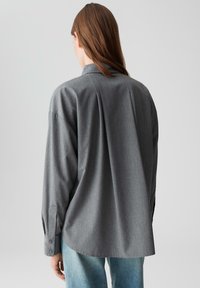 Woman with long brown hair wearing a loose gray long-sleeve shirt and light blue jeans, standing facing away against a plain gray background.