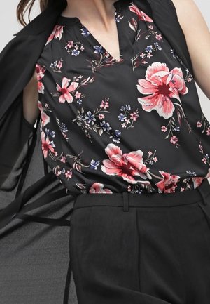 Vrouw die een zwarte mouwloze blouse draagt met een roze en rode bloemenprint, gecombineerd met een zwarte hooggetailleerde broek en een vloeiende zwarte jas.