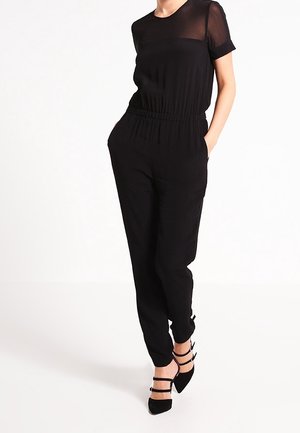 Czarny kombinezon z krótkimi przezroczystymi rękawami i marszczoną talią. Posiada zwężane nogawki i boczne kieszenie. Zestawiony z czarnymi sandałkami na paski.