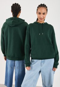 Grønn hettegenser laget av mykt stoff. Har en frontlomme, justerbare snorer og ribbestrikkede ermer. Brukt med lyseblå jeans.