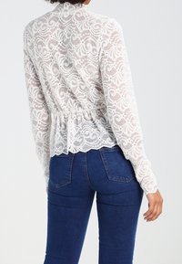 Blouse en dentelle blanche avec des motifs floraux complexes, manches longues et taille froncée. Portée sur un jean en denim foncé.