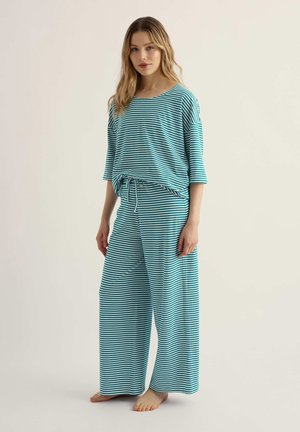 Žena stojaca na boso, oblečená v zodpovedajúcom voľnom topu a širokých nohaviciach s horizontálnymi pruhmi v tyrkysovej a bielej farbe, s nastaviteľným pásom na šnúrku.