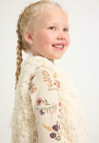 Faux-Fur-Weste in Elfenbein über einem gerippten, langärmeligen Oberteil mit floralen Mustern in Rosatönen, Lila und Grün. Blonde, geflochtene Haare.
