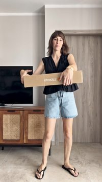 Kartonska kutija u rukama, označena "Zalando", svijetlosmeđe boje. Na sebi jednostavna crna bluzica, traper šorts i crne japanke.