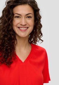 Czerwona bluzka z dekoltem w serek i krótkimi rękawami, z falbaniastym wykończeniem przy dekolcie oraz miękką, gładką fakturą. Modelka ma kręcone włosy i przyjazny wyraz twarzy.