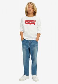 Hosszú ujjú fehér ing piros Levi's logóval, kék farmernadrággal és fehér tornacipővel párosítva. A szabás kényelmes, klasszikus kialakítású.