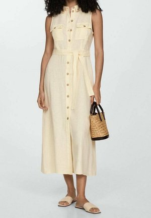 Vrouw draagt een mouwloze beige midi-jurk met knopen aan de voorkant en een riem, houdt een gevlochten stro-handtas vast en draagt beige sandalen zonder achterkant.