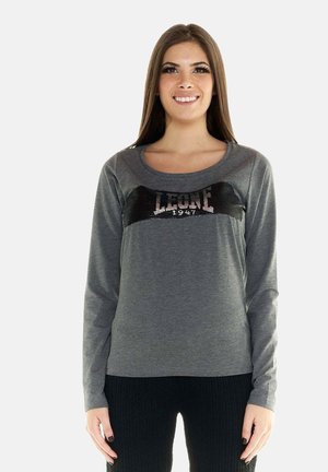 Donna sorridente con lunghi capelli castani che indossa una maglia grigia a maniche lunghe con testo nero in paillettes "LEONE 1947" e pantaloni neri strutturati.