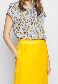 Blusa azzurro chiaro a maniche corte con motivo floreale marrone, con scollatura allacciata. Abbinata a una gonna gialla a vita alta con tasche.