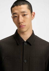Brown herringbone shirt jacket featuring a collar, two chest pockets, and button closures. The fabric has a textured, woven appearance.