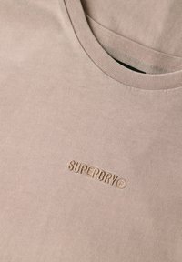 Lyse beige bomulds t-shirt med ribbet halsudskæring og prægede brandlogo i matchende farve på den nederste front. Glat tekstur.