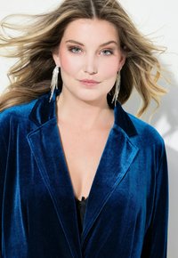 Samet bleiseri syvässä sinisessä värissä, jossa on shuolikaulus. Mallilla on pitkät, kimaltavat korvakorut. Pehmeä rakenne ja elegantti malli näkyvät.