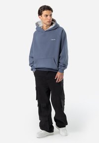 Pulover cu glugă albastru, cu buzunar frontal și logo alb, asortat cu pantaloni cargo negri. Modelul poartă adidași albi.