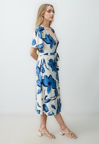 Blauwe bloemenprint wikkeljurk met korte fladdermouwen en een strik taille, met een V-hals en een middenlengte zoom. Gepaard met witte blokhak sandalen.