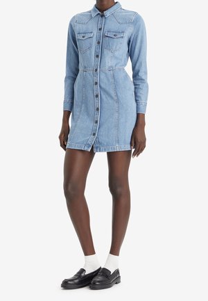 Woman wearing a light blue denim button-up dress with long sleeves, front pockets, white socks, and black loafers, standing against white background.