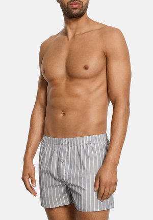 Mężczyzna bez koszulki z brodą, ubrany w szare i białe bokserki w pionowe paski z elastycznym pasem, na białym tle.