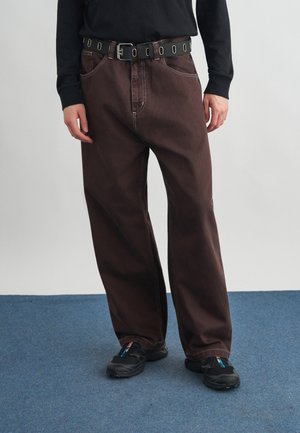 Άτομο που φοράει φαρδιά σκούρα καφέ παντελόνια με μαύρη ζώνη, μαύρο μακρυμάνικο τοπ και μαύρα αθλητικά παπούτσια, στεκεμένο πάνω σε μπλε χαλί.